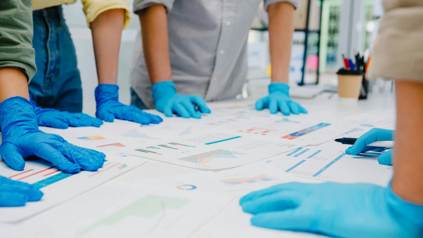 A team in blue nitrile gloves examining bar charts and printed data sheets across a table, the kind of comparative review used in surveillance.