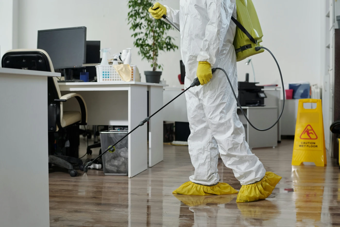 A worker in a white Tyvek suit and yellow gloves spraying disinfectant in an indoor space, the kind of cleanup that creates aerosol exposure if done dry.