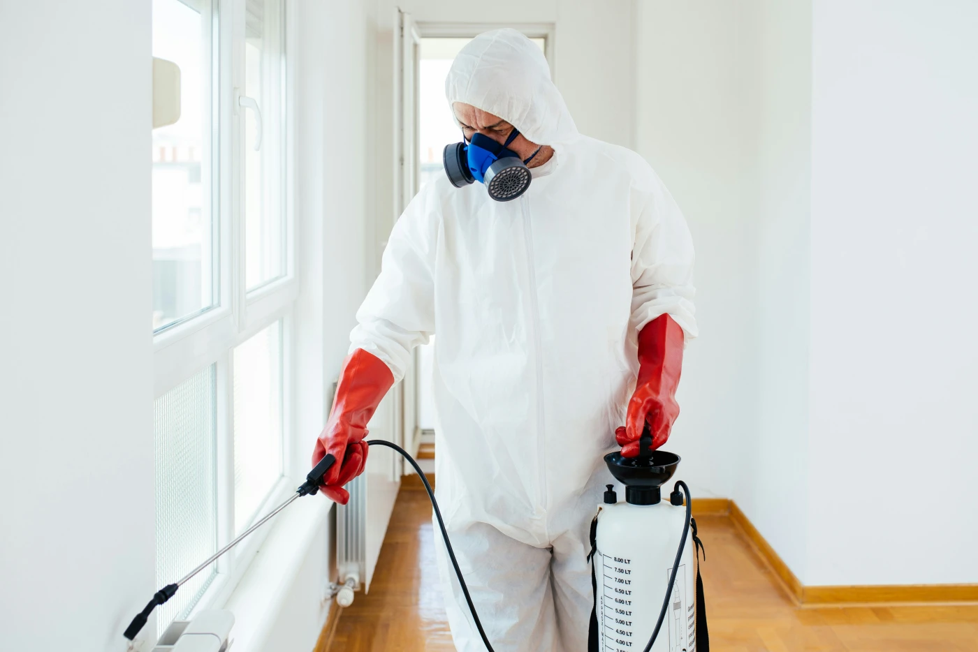 A pest-control technician in a Tyvek suit, respirator, and red gloves treating a residential interior with a pump sprayer.