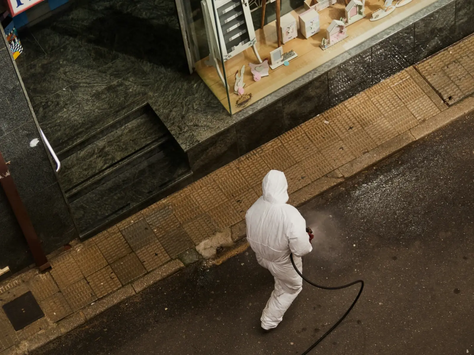 A worker in a white protective suit walking past a building entrance with a chemical sprayer, the kind of contractor used for outdoor and structural cleanup.